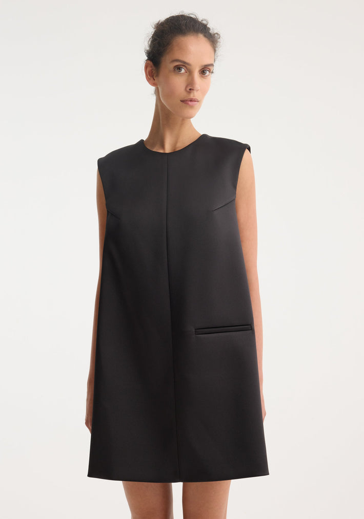 Model in Sculptural cape dress | black, sleeveless with round neckline, straight silhouette, above-knee length, featuring a welt pocket and dramatic back box pleat.