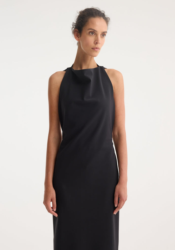 Open back dress | black: Woman in a sleeveless, floor-length black dress with a high draped neckline, open back, and tie detail, posing slightly sideways.