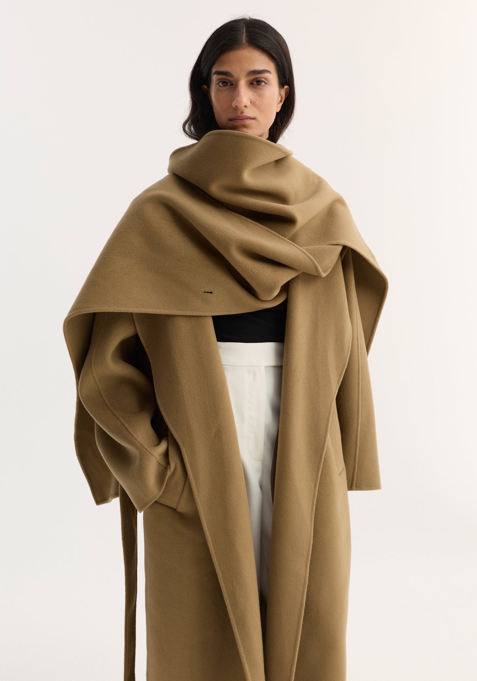 Woman wearing a camel Signature double-faced scarf coat | camel with a large draped scarf collar over a black top and white pants against a plain background.