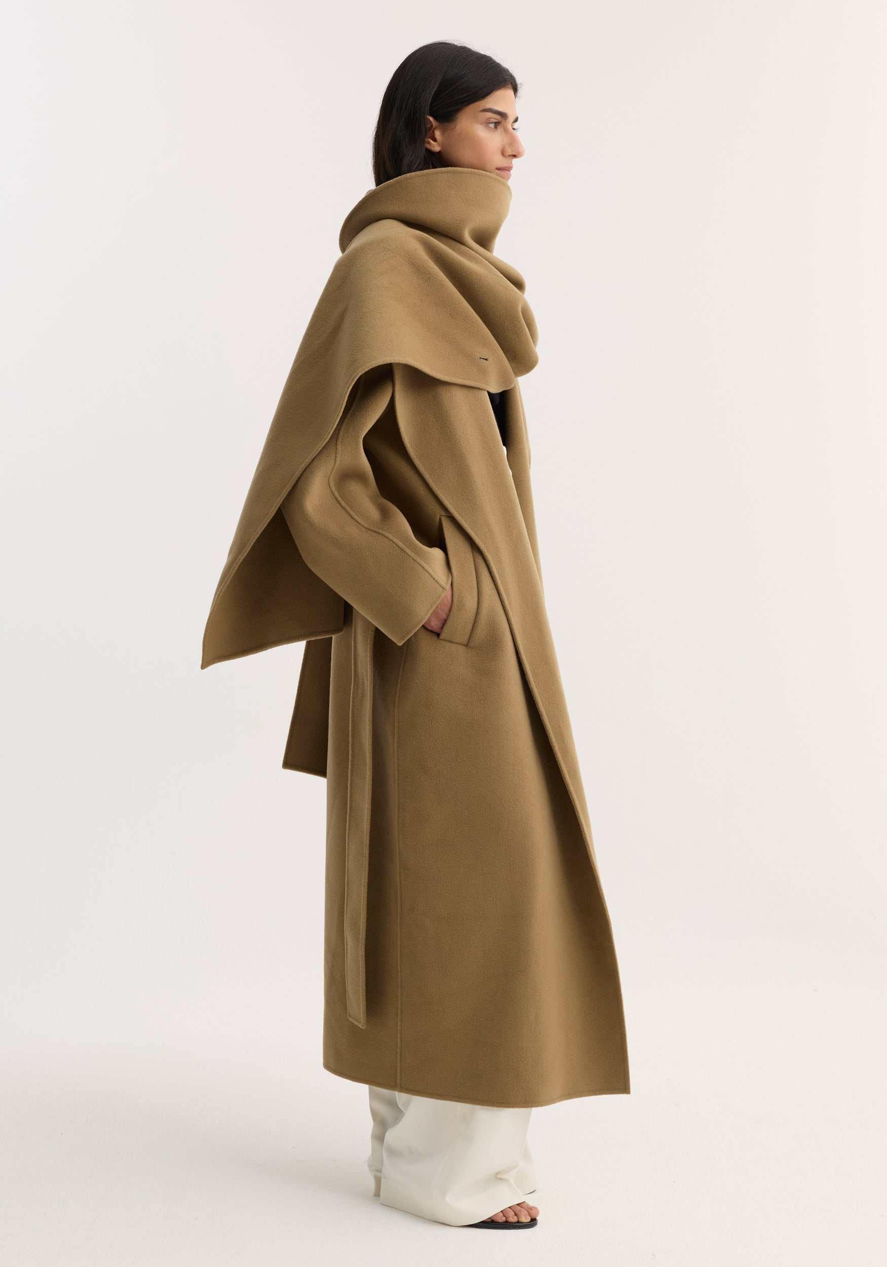 Woman wearing a camel-colored Signature double-faced scarf coat with a high, draped collar and long sleeves, styled with white wide-leg pants against a plain background.