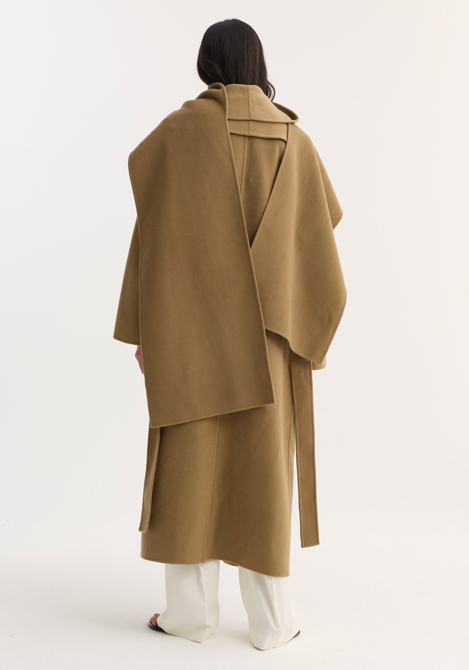 Back view of a person wearing a camel-colored Signature double-faced scarf coat | camel with a long scarf detail draped over the shoulders, paired with white pants.