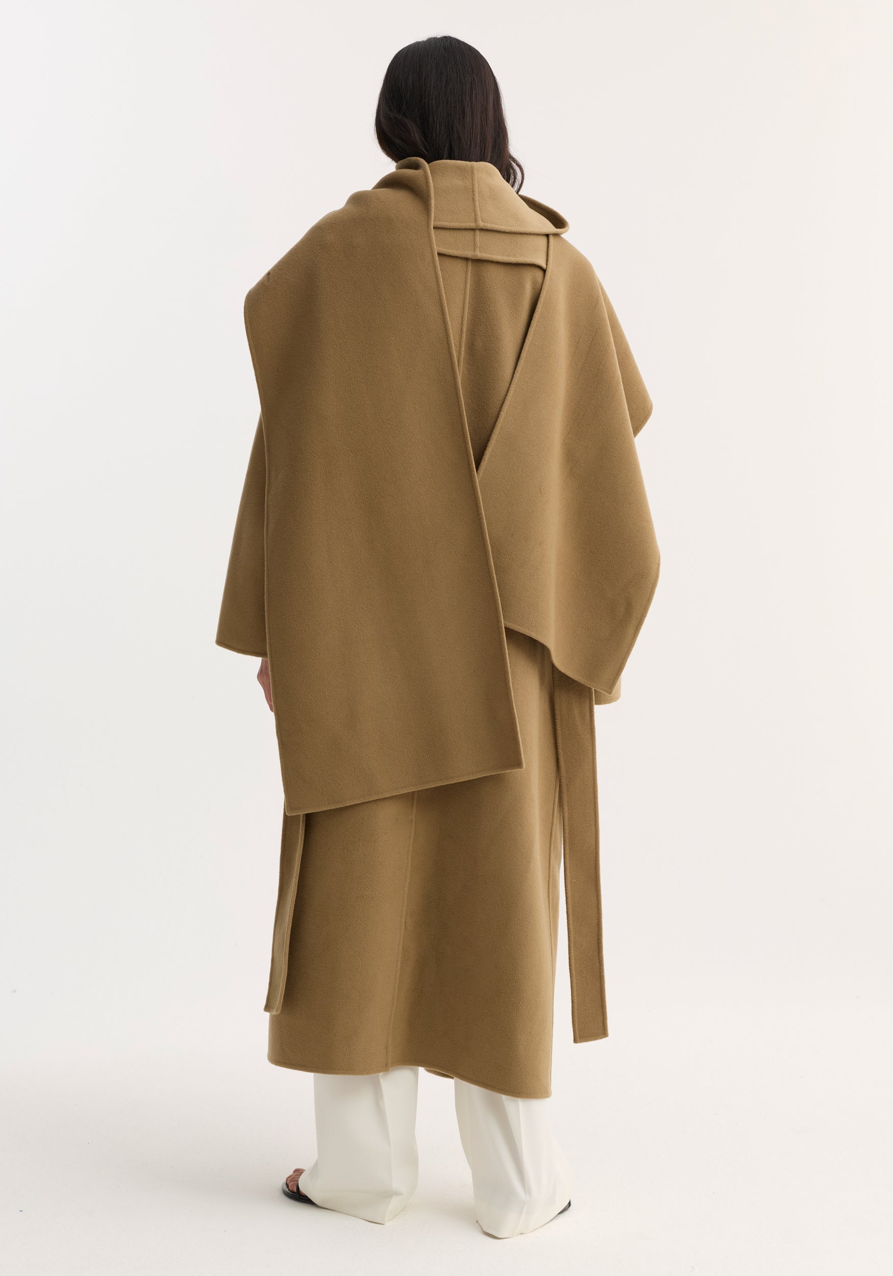 Back view of a person wearing a camel-colored Signature double-faced scarf coat | camel with a long scarf detail draped over the shoulders, paired with white pants.