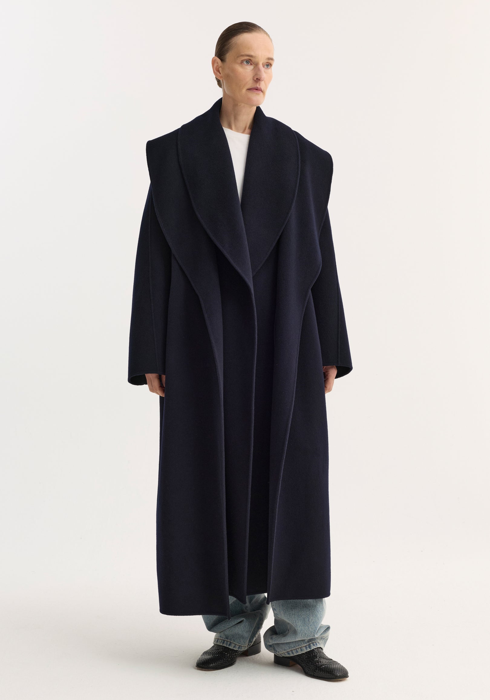 Woman wearing a long navy Signature double-faced scarf coat | navy with wide lapels over casual jeans and black shoes against a plain white background.