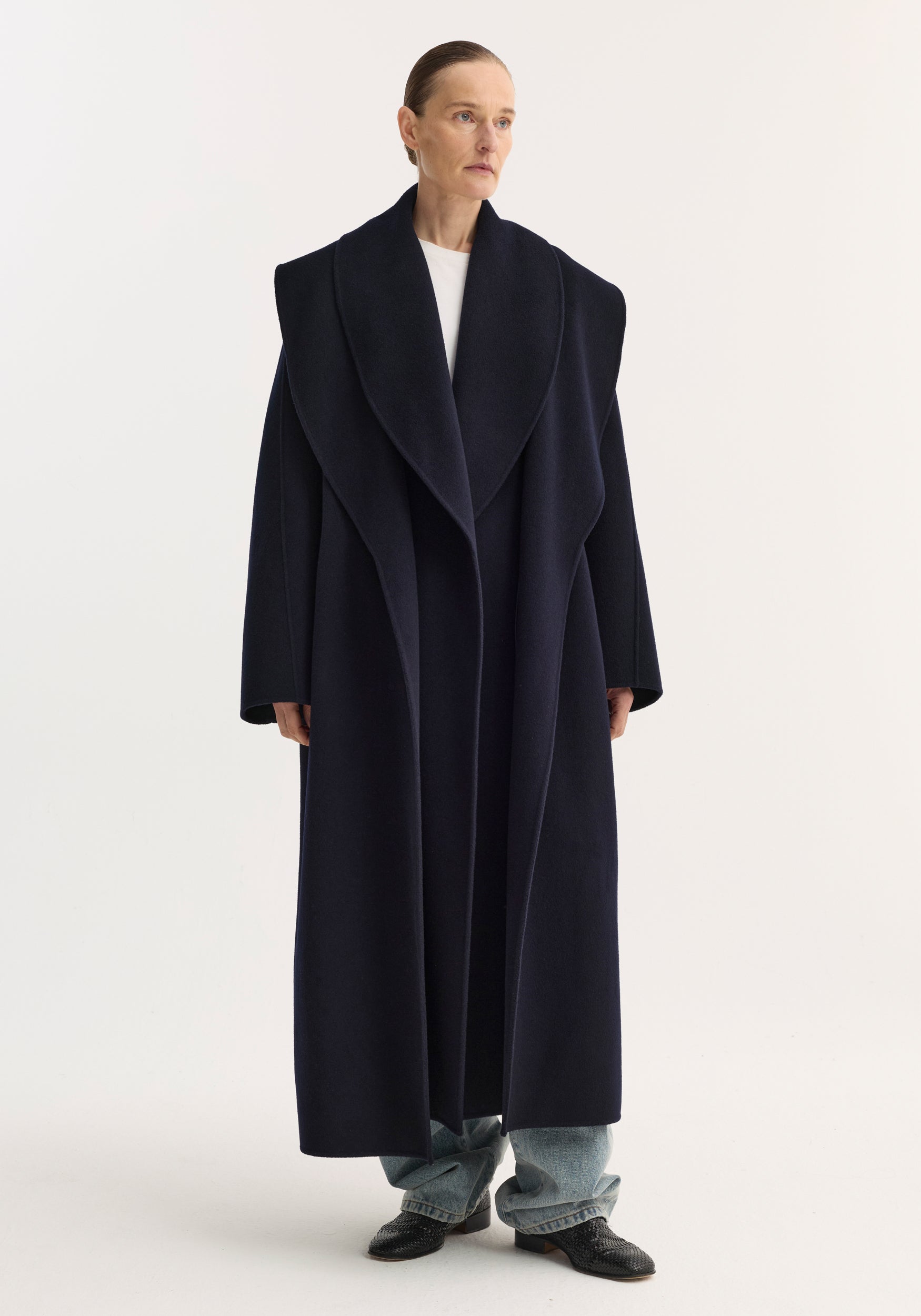 Woman wearing a long navy Signature double-faced scarf coat | navy with wide lapels over casual jeans and black shoes against a plain white background.