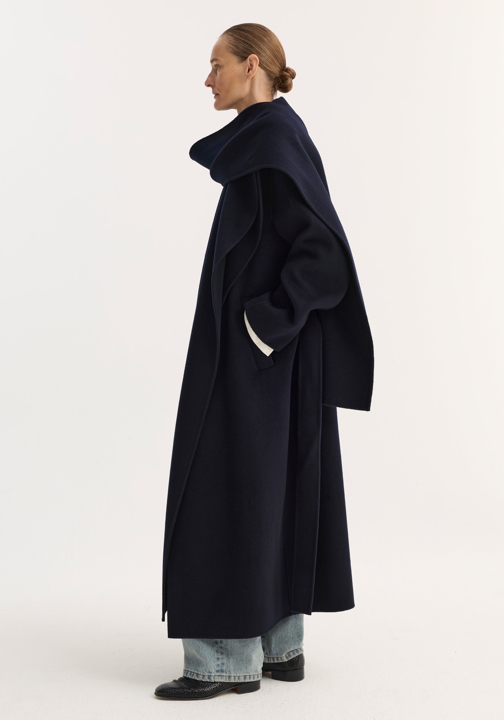 Side view of a woman wearing a long navy coat with a large attached scarf draped over her shoulders, styled with light blue jeans and black shoes, featuring the Signature double-faced scarf coat | navy.
