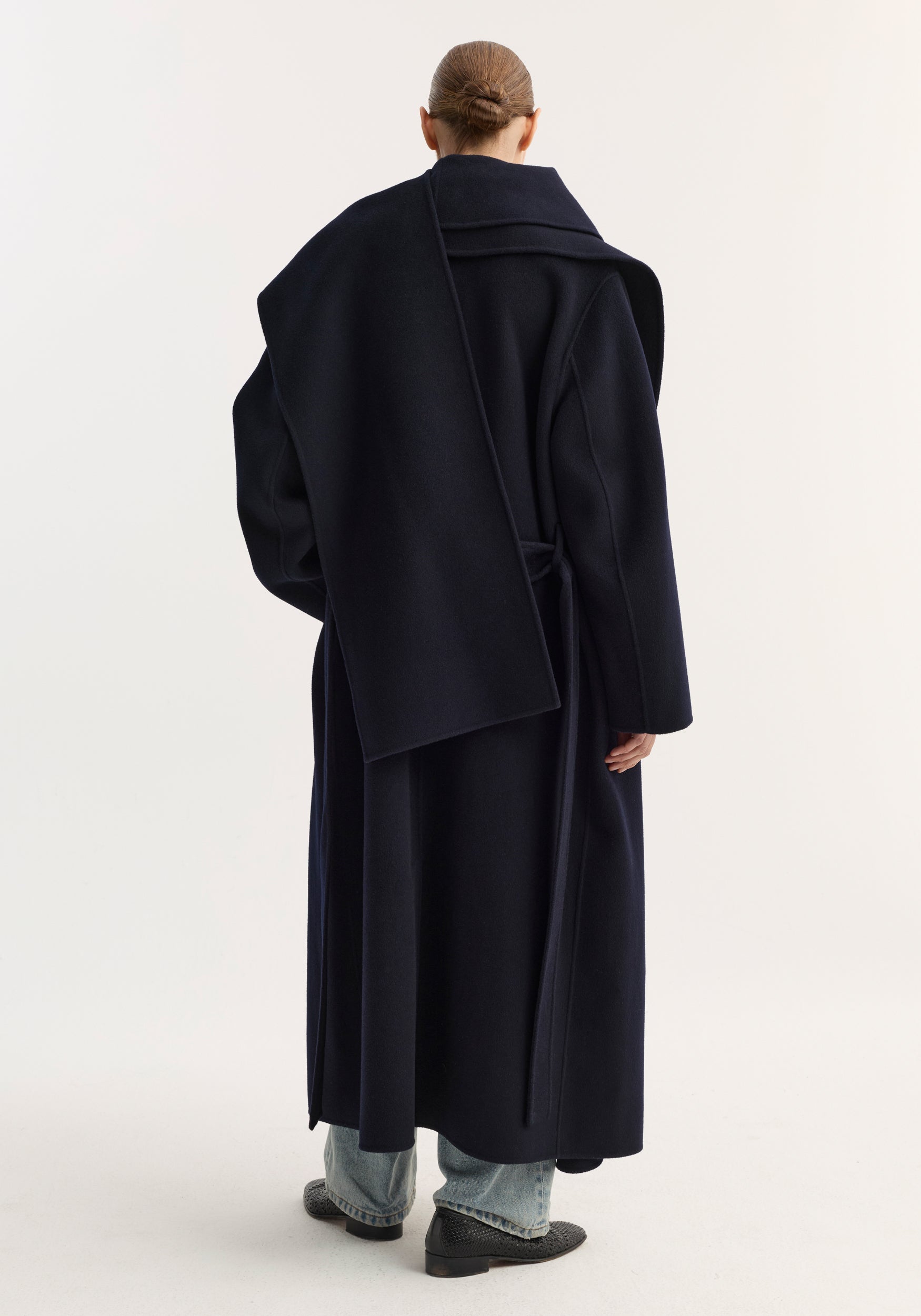 Woman seen from the back wearing a navy long signature double-faced scarf coat | navy with a wide scarf detail draped over one shoulder, paired with light blue jeans and black shoes.
