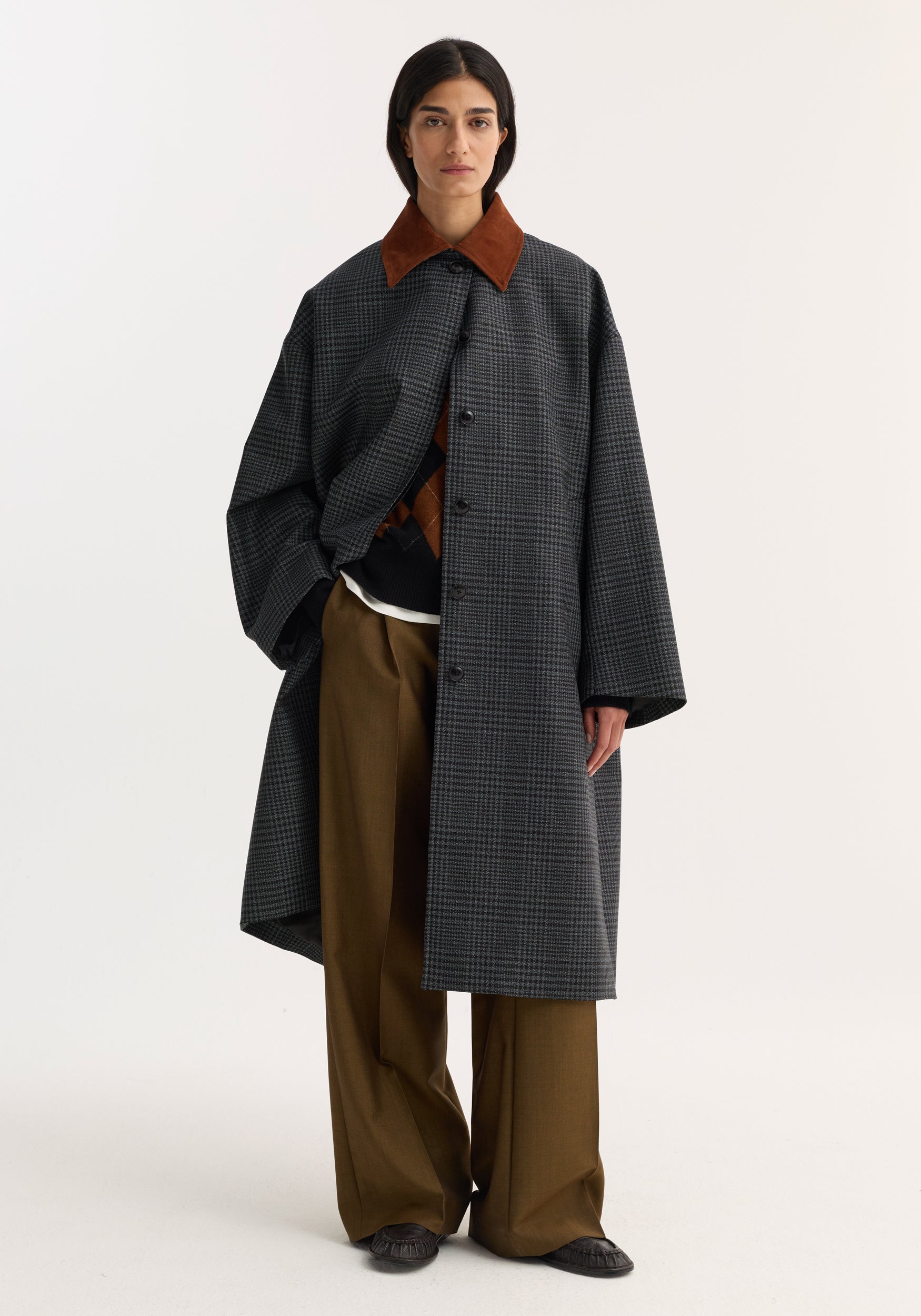 A model wearing a long Suede collar check coat | black / grey check with a brown suede collar, styled over wide-leg brown trousers and standing against a plain light background.