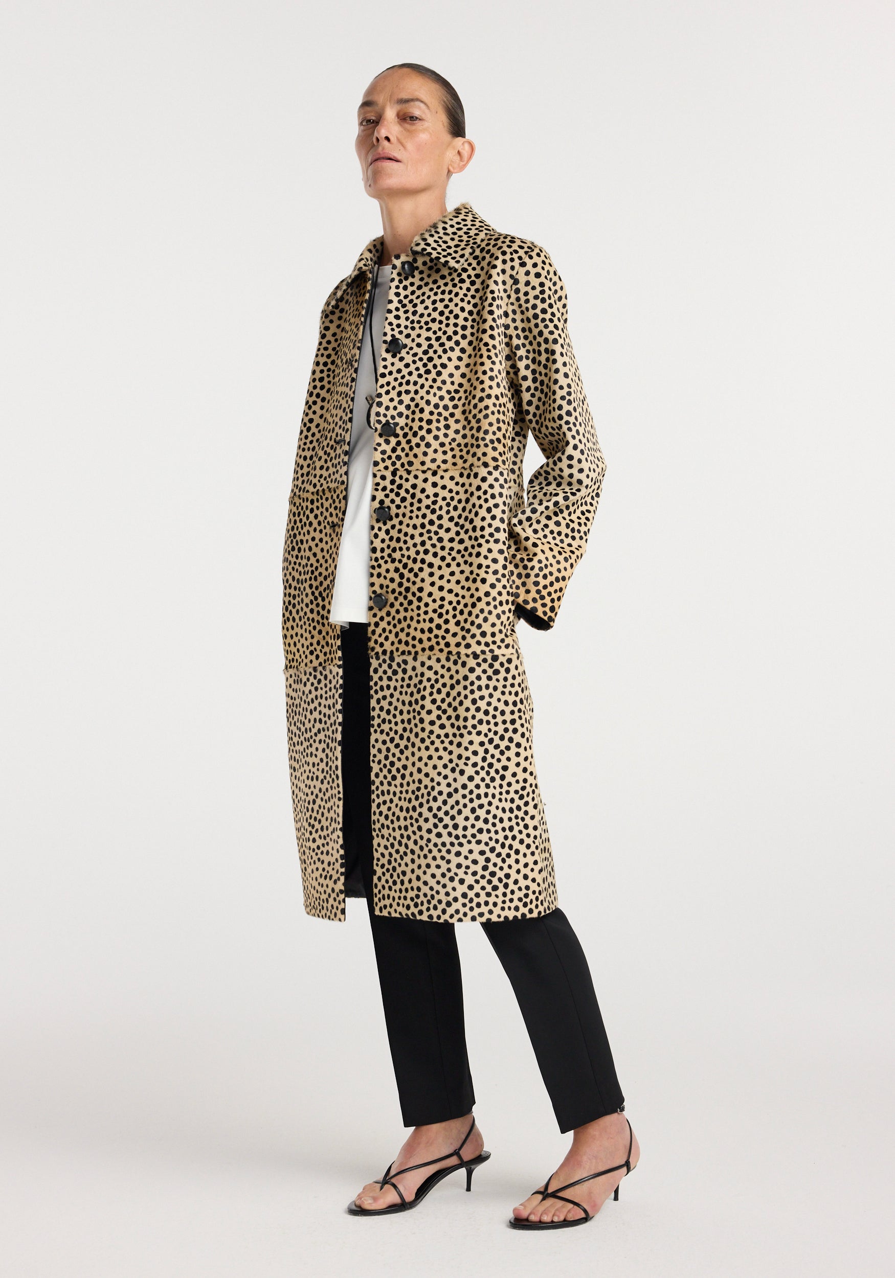 Woman wearing a long cheetah print Pony hair coat | cheetah with button closure, paired with black pants and strappy black heels against a plain background.