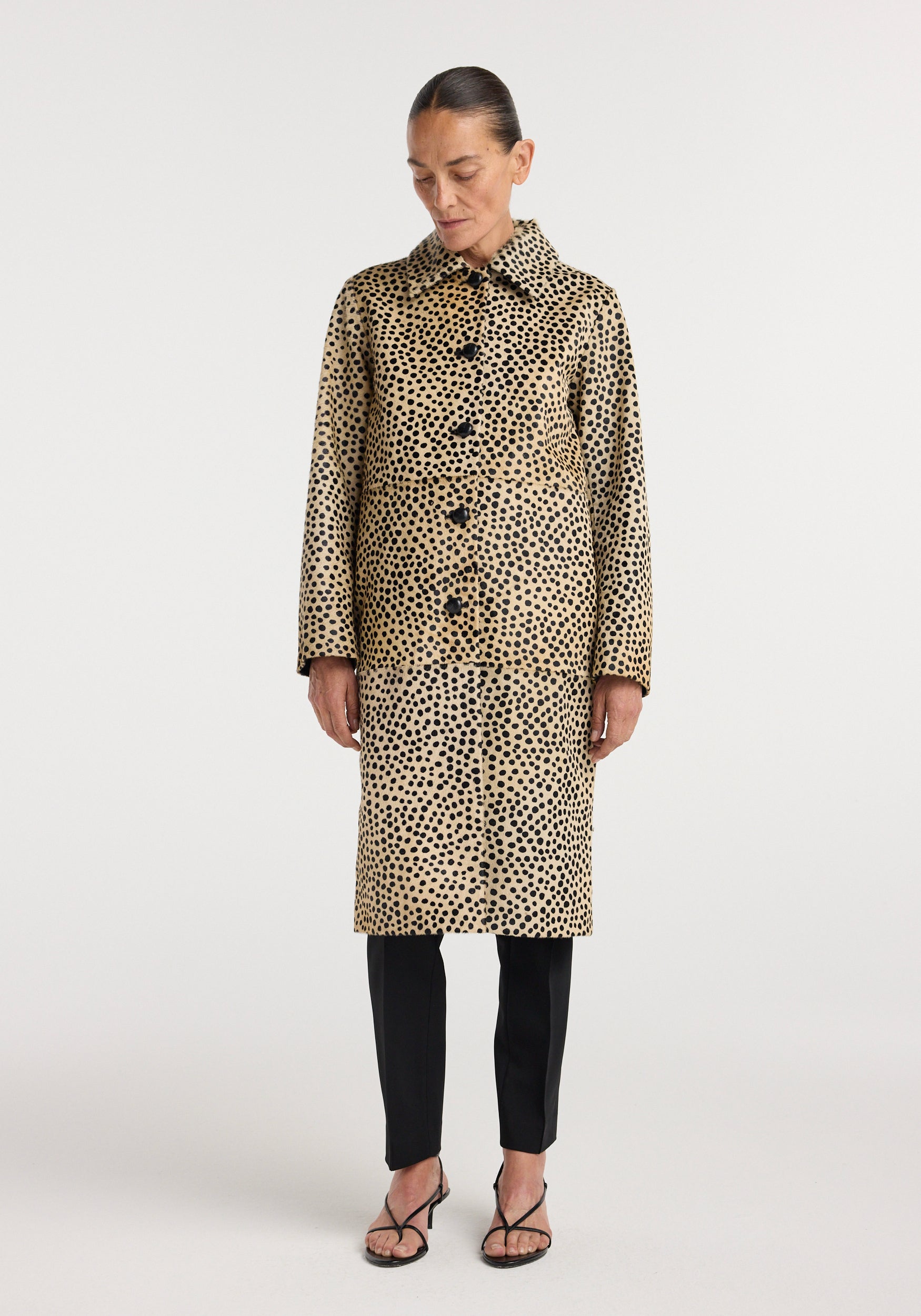 Woman wearing a long cheetah print pony hair coat | cheetah with black buttons, paired with black pants and strappy black sandals, standing against a plain background.