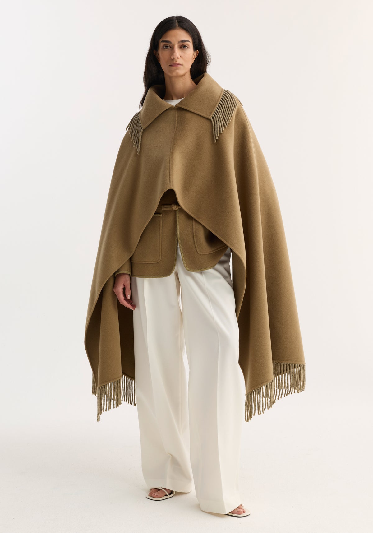 Woman wearing a camel-colored Fringe scarf poncho with fringe details on the collar and hem, paired with white wide-leg trousers and an olive green jacket underneath.