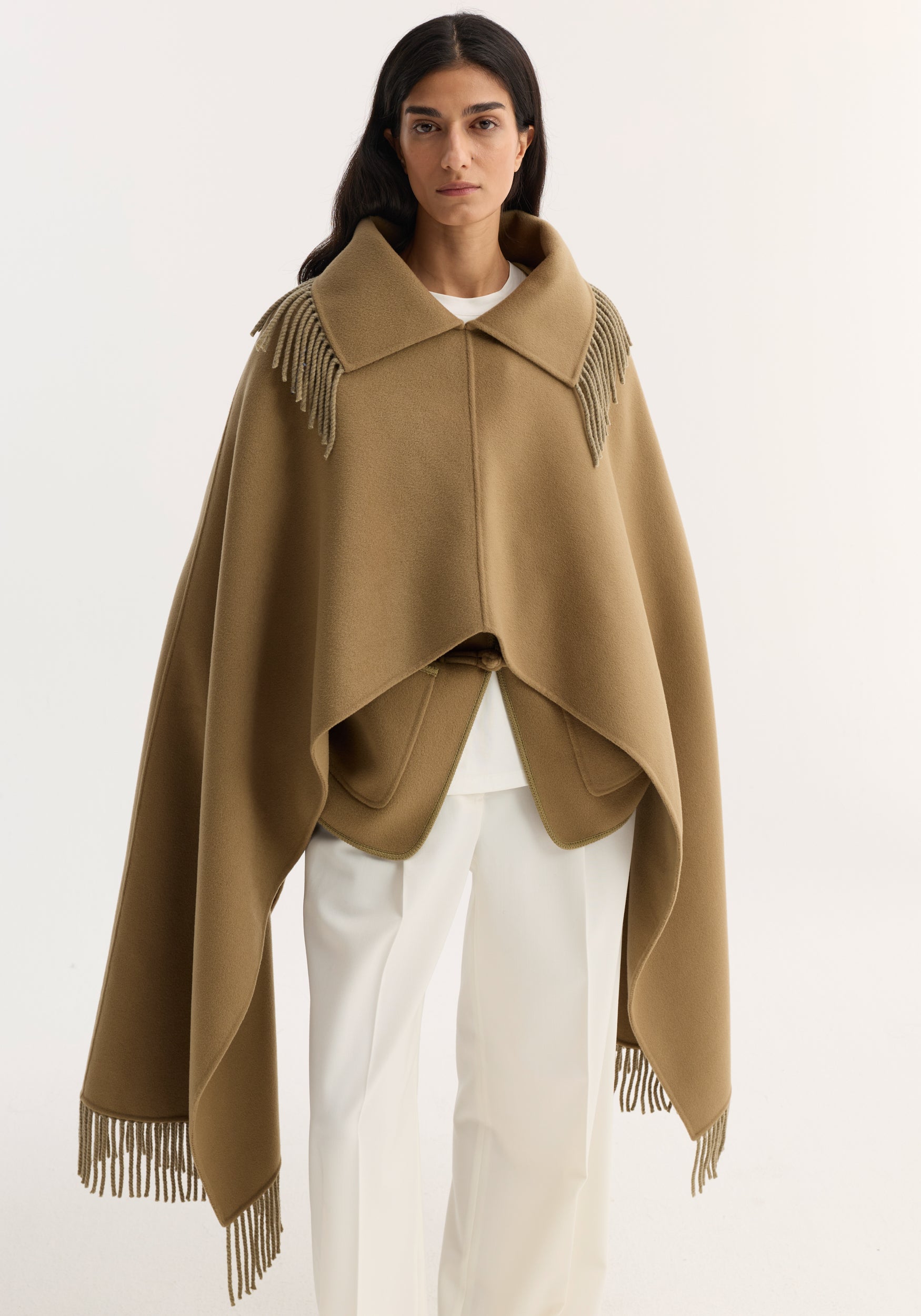 Woman wearing a camel-colored Fringe scarf poncho with wide collar, draped over white pants, featuring fringe details along the shoulders and hem.