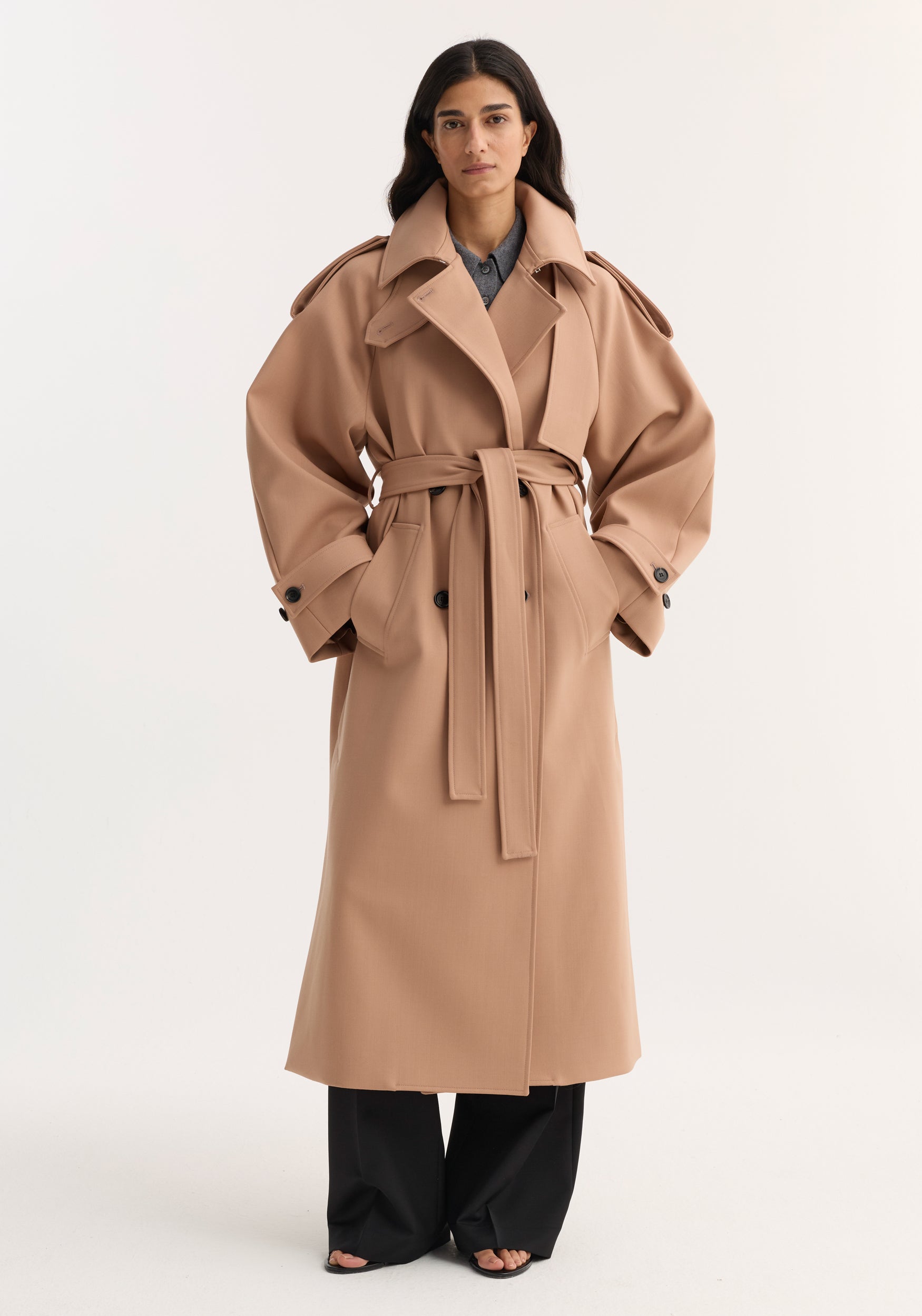 A woman wearing an oversized classic trench | powder in a light beige tone with wide sleeves, a belted waist, large lapels, and buttoned cuffs, styled over black wide-leg pants.