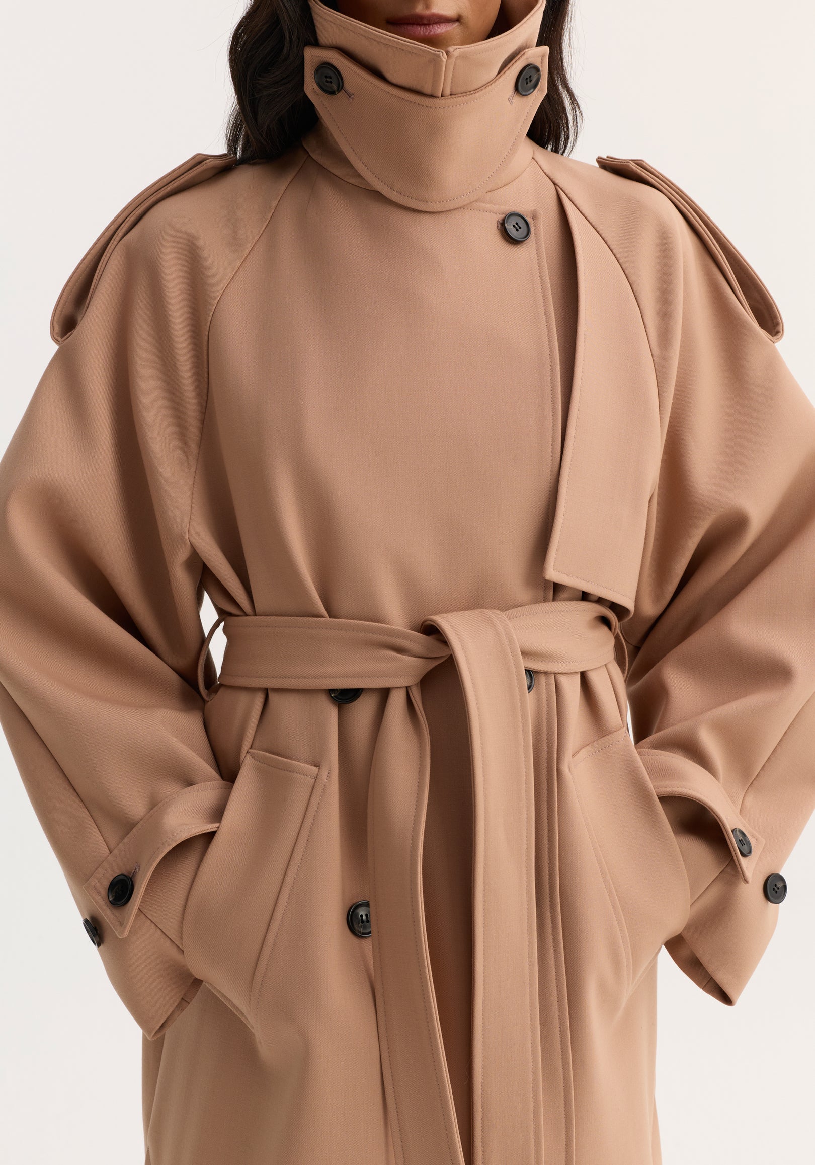 Close-up of a person wearing an oversized classic trench | powder with a high collar, button details, wide sleeves, and a waist belt tied in the front.