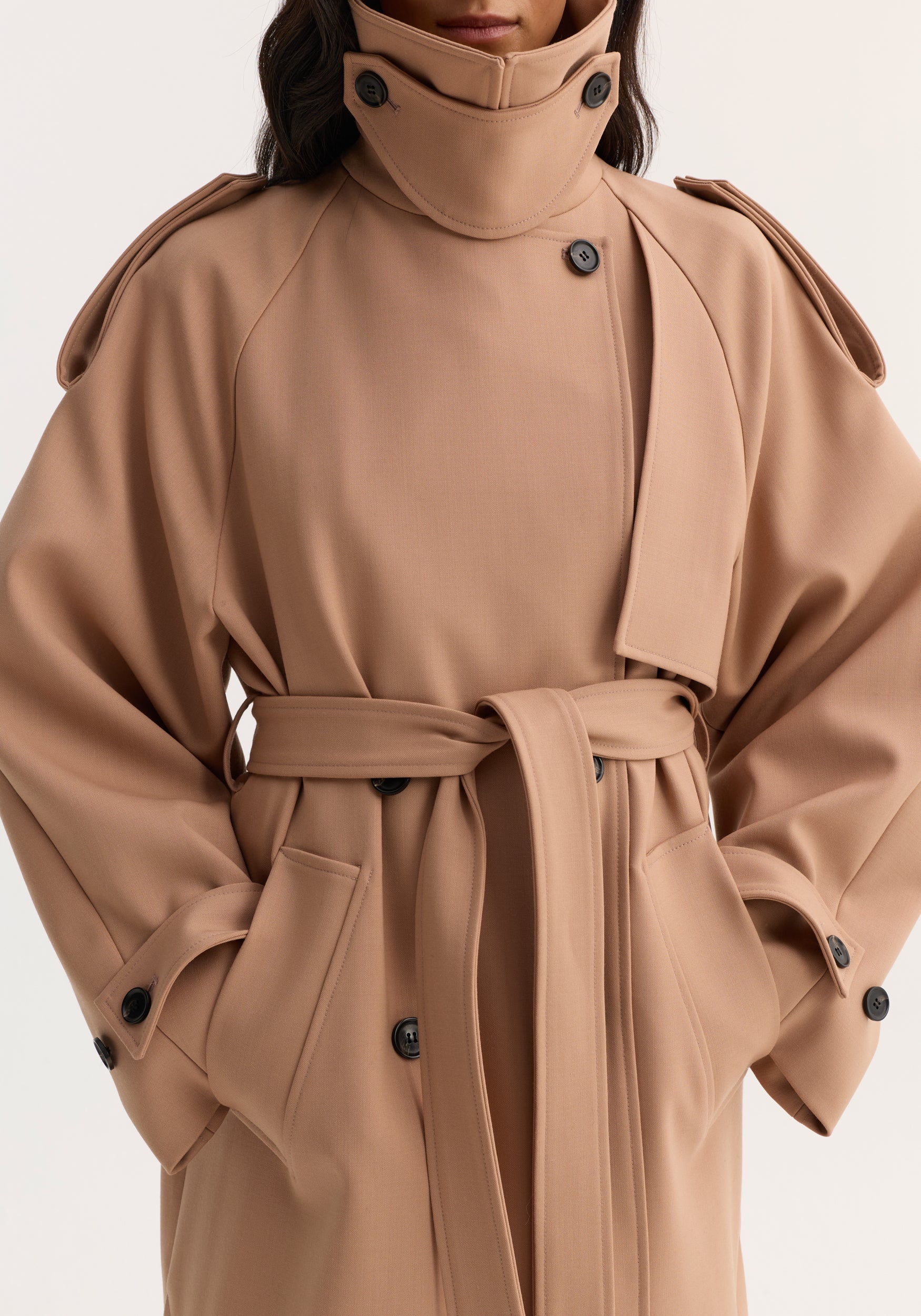Close-up of a person wearing an oversized classic trench | powder with a high collar, button details, wide sleeves, and a waist belt tied in the front.