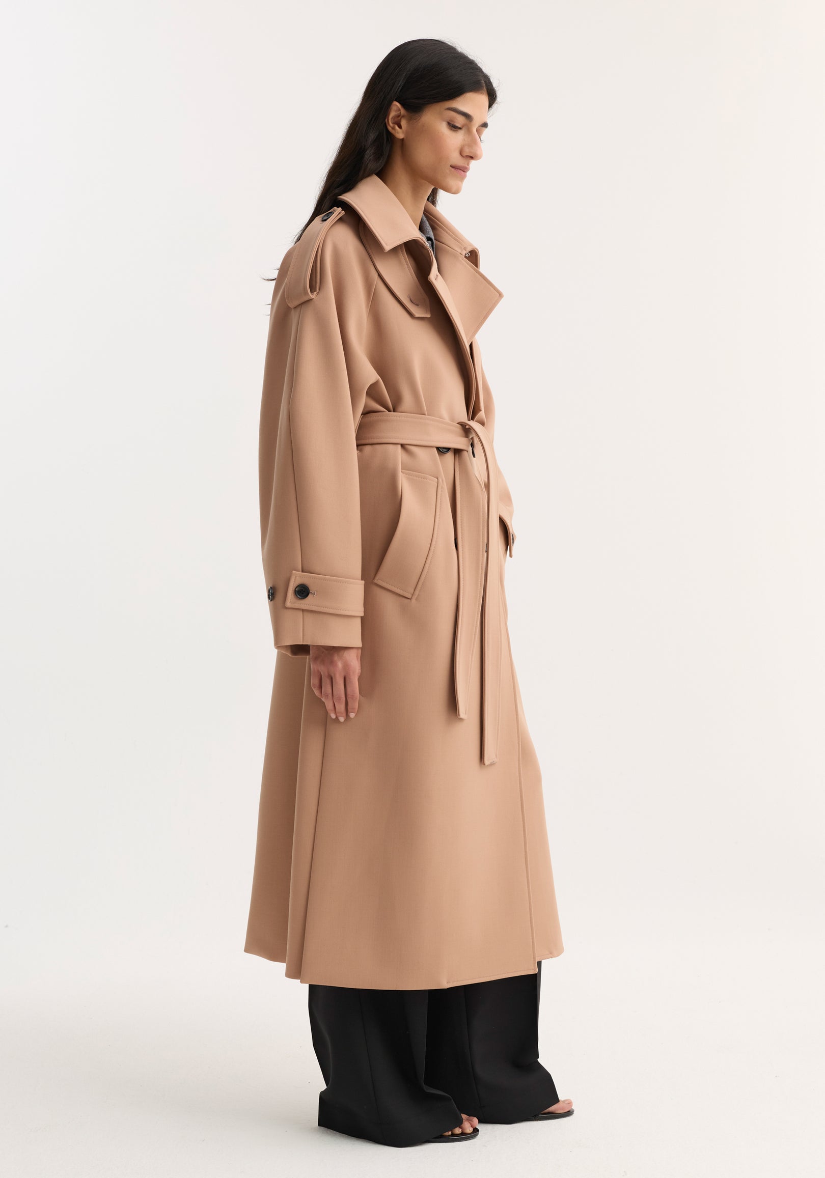 Woman wearing an oversized classic trench coat in powder color, featuring a belted waist, wide lapels, and buttoned cuffs, styled with black pants and open-toe shoes.