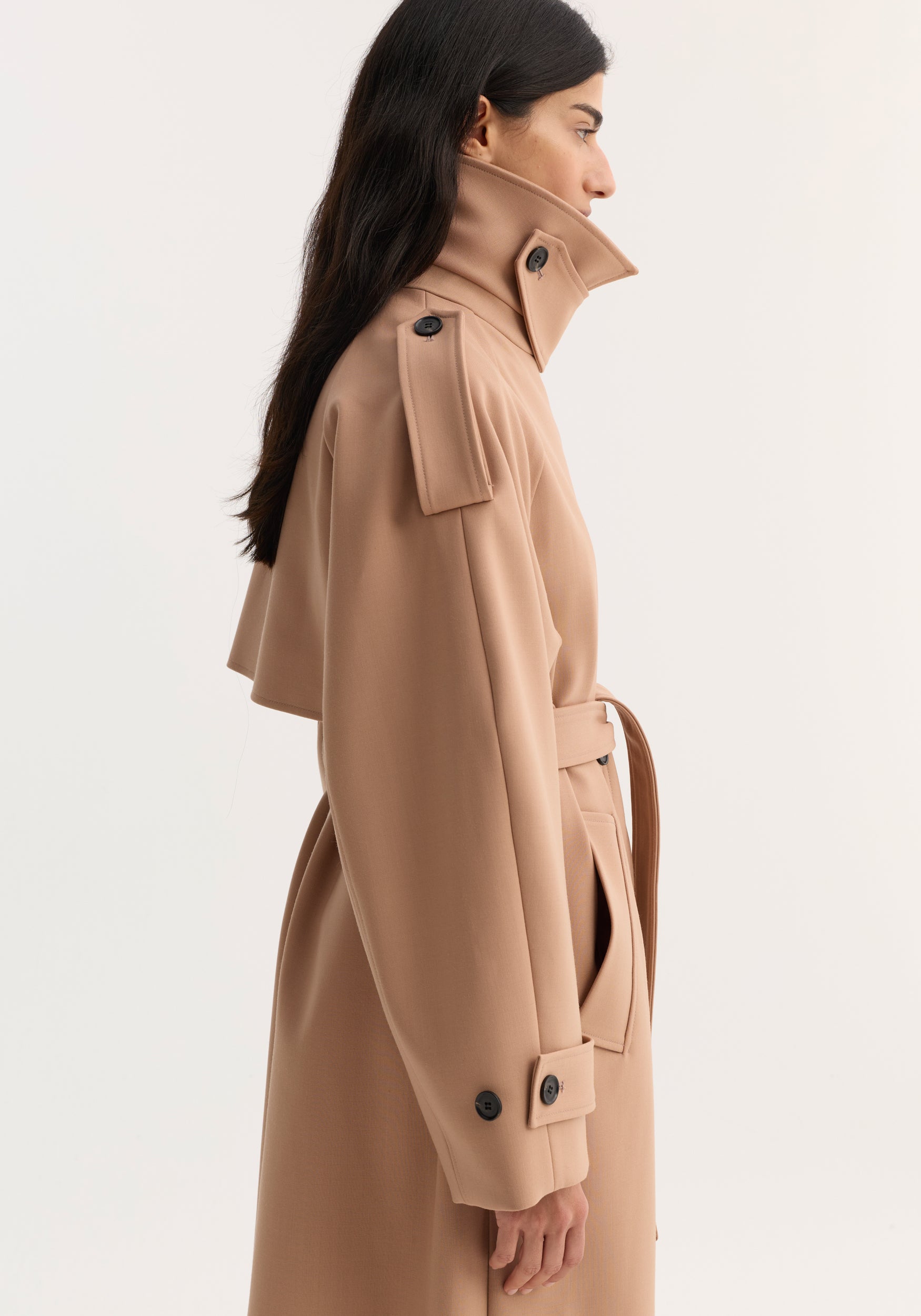 Side view of a woman wearing an oversized classic trench | powder with a high collar, shoulder epaulets, sleeve straps, belt cinching the waist, and a smooth fabric finish.