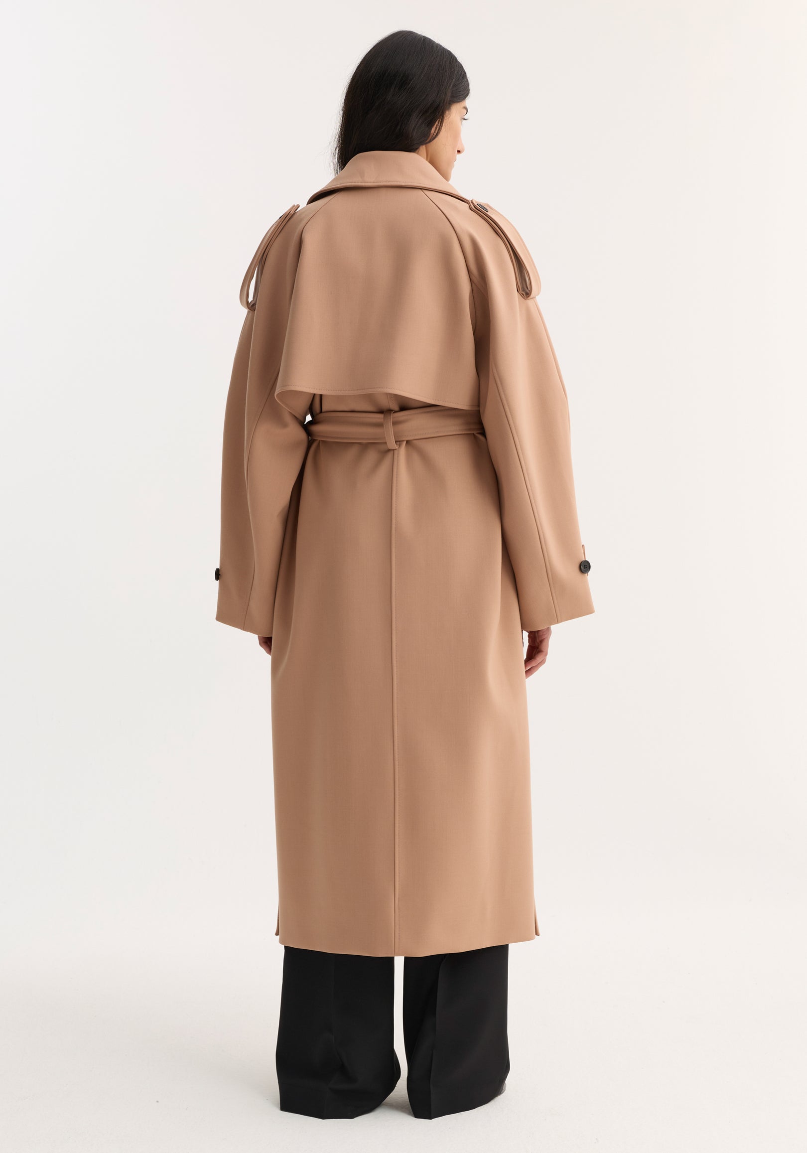 Back view of a woman wearing an oversized classic trench | powder in a soft beige tone, featuring a belt at the waist and buttoned cuffs, paired with black wide-leg pants.