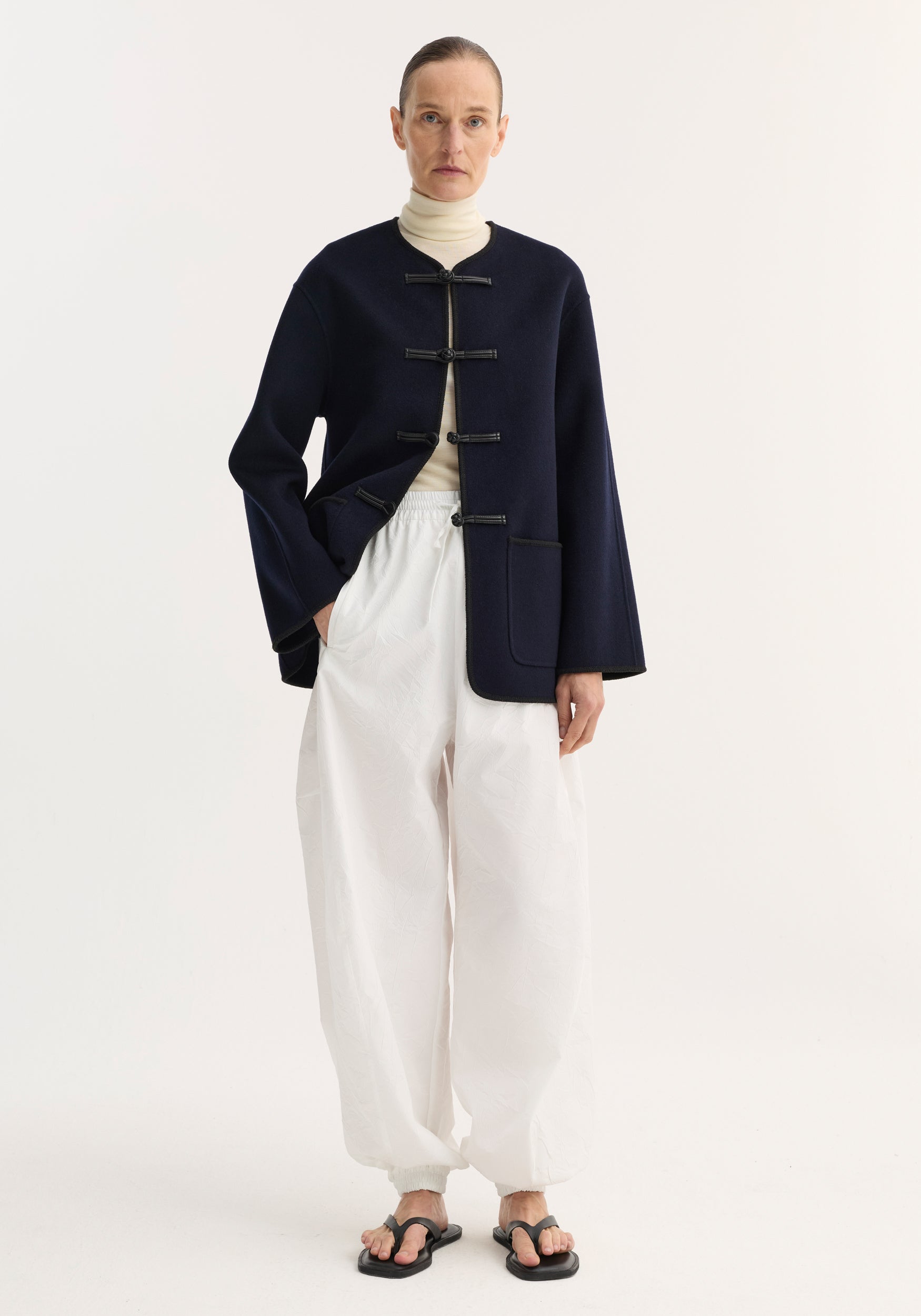 A model wearing a navy Double-faced wool pankou jacket | navy over a cream turtleneck, paired with loose white pants and black sandals, standing against a plain white background.
