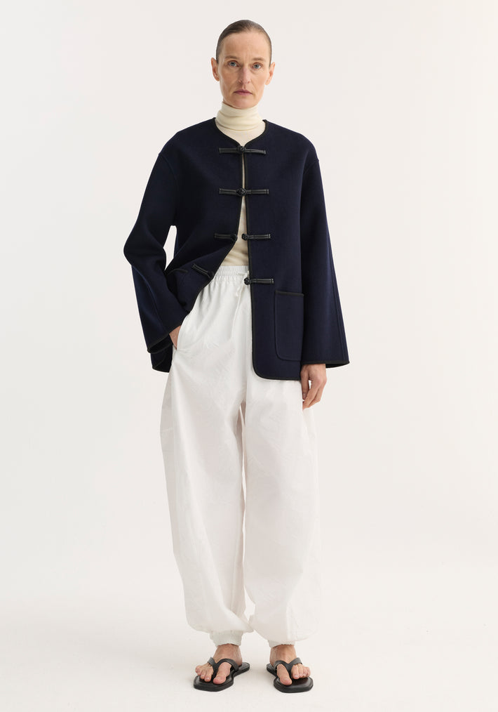 Model wearing Double-faced wool pankou jacket in navy with pankou knot closures, over cream turtleneck, paired with white trousers and black sandals.