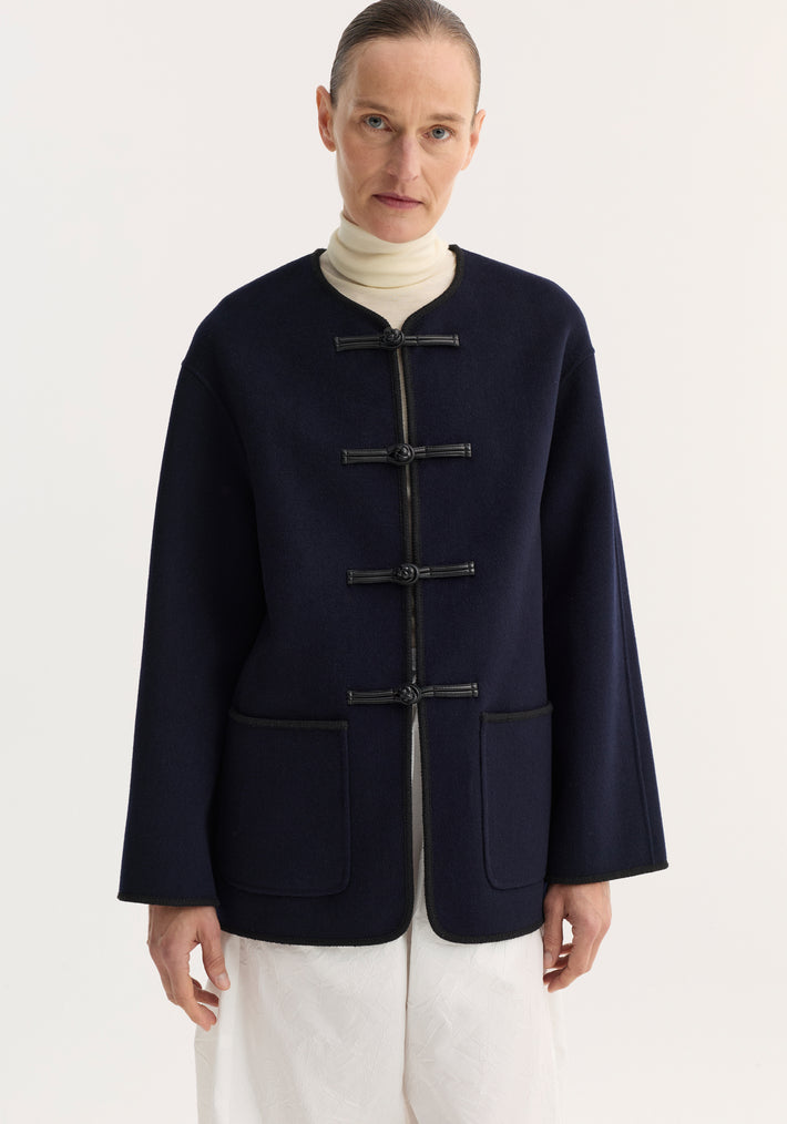 Model wearing a Double-faced wool pankou jacket in navy with frog-style closures, patch pockets, black piped edges, paired with cream top and white trousers.