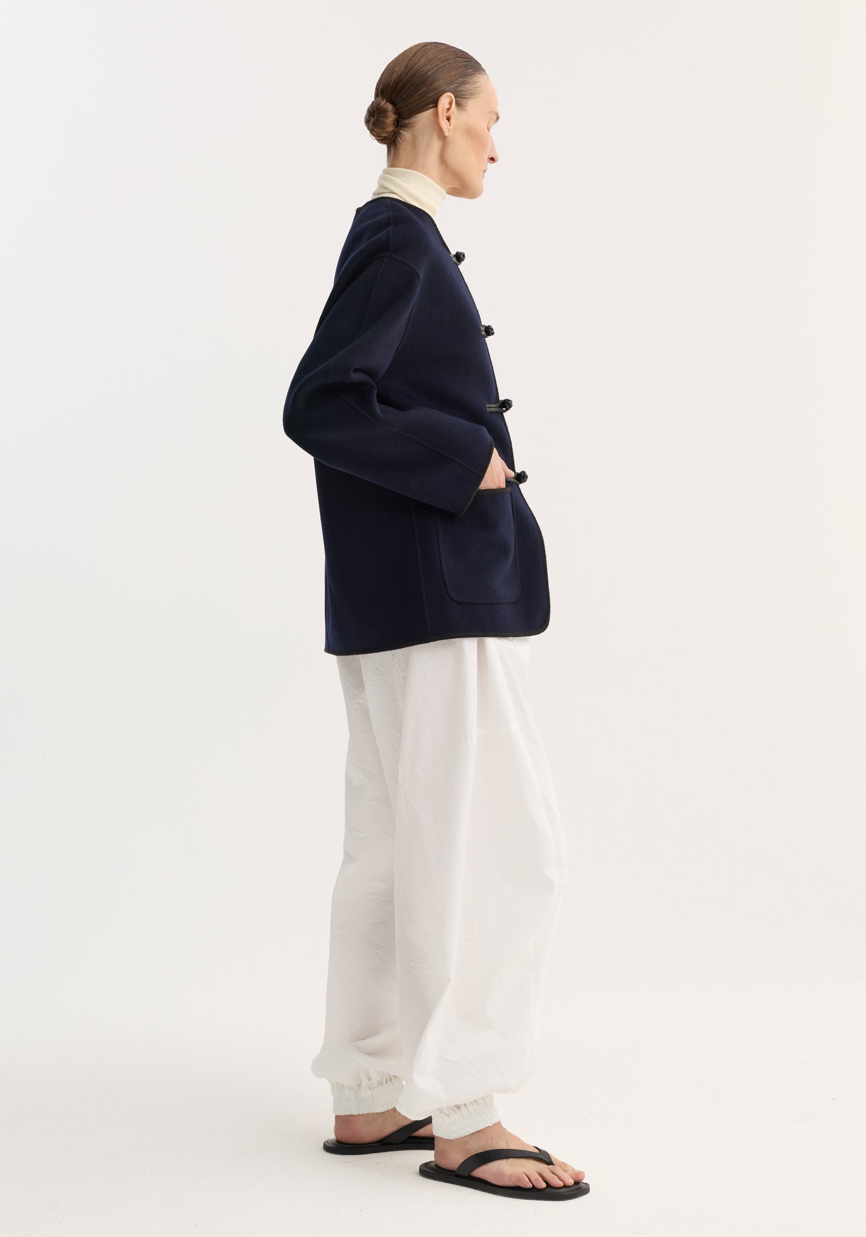 Woman wearing a navy Double-faced wool pankou jacket with frog button closures, paired with white loose pants and black sandals, standing in profile against a plain background.