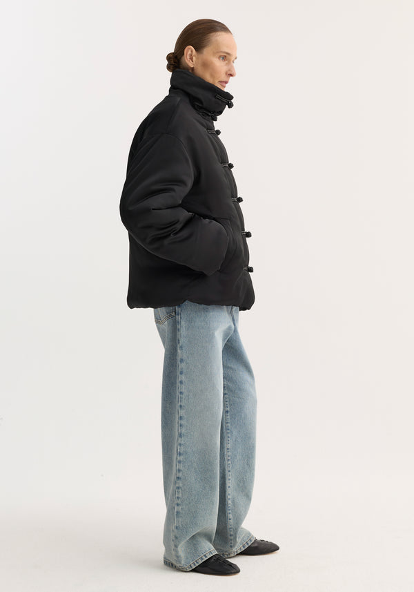 Side-profile of a model in a Padded puffer jacket | black, featuring a high collar, pankou closures, and slanted welt pockets, paired with wide-leg jeans.
