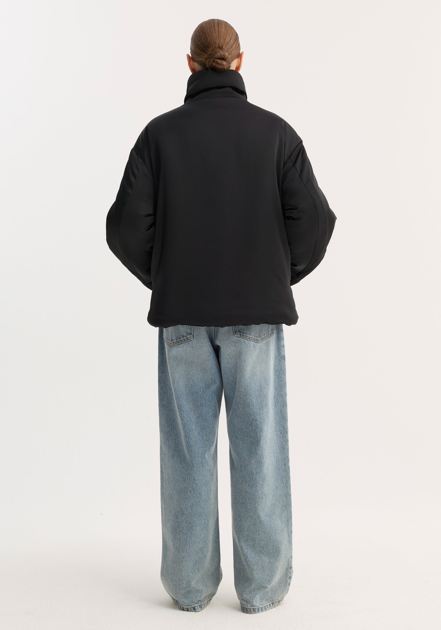 Back view of a person wearing a black padded puffer jacket | black with a high collar and loose fit, paired with light blue wide-leg jeans against a plain background.