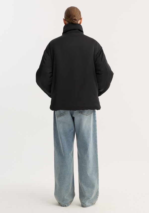 A person models a Padded puffer jacket | black, showcasing its boxy silhouette, high collar, and pankou closures, paired with wide-leg jeans.