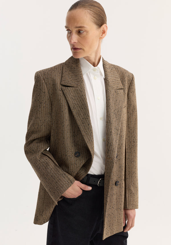 Double breasted blazer | herringbone: Person in a herringbone-textured double-breasted blazer with chest and welt pockets, over a white shirt, hand in trouser pocket.