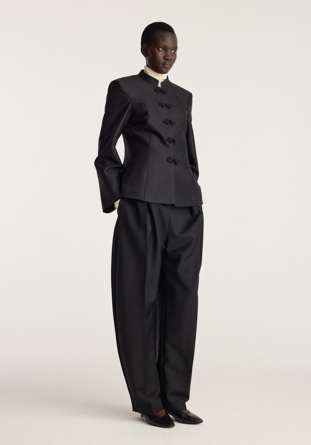 Sculptural pankou closure blazer | black worn by a model with wide leg black trousers and pointed shoes in a minimalist studio setting
