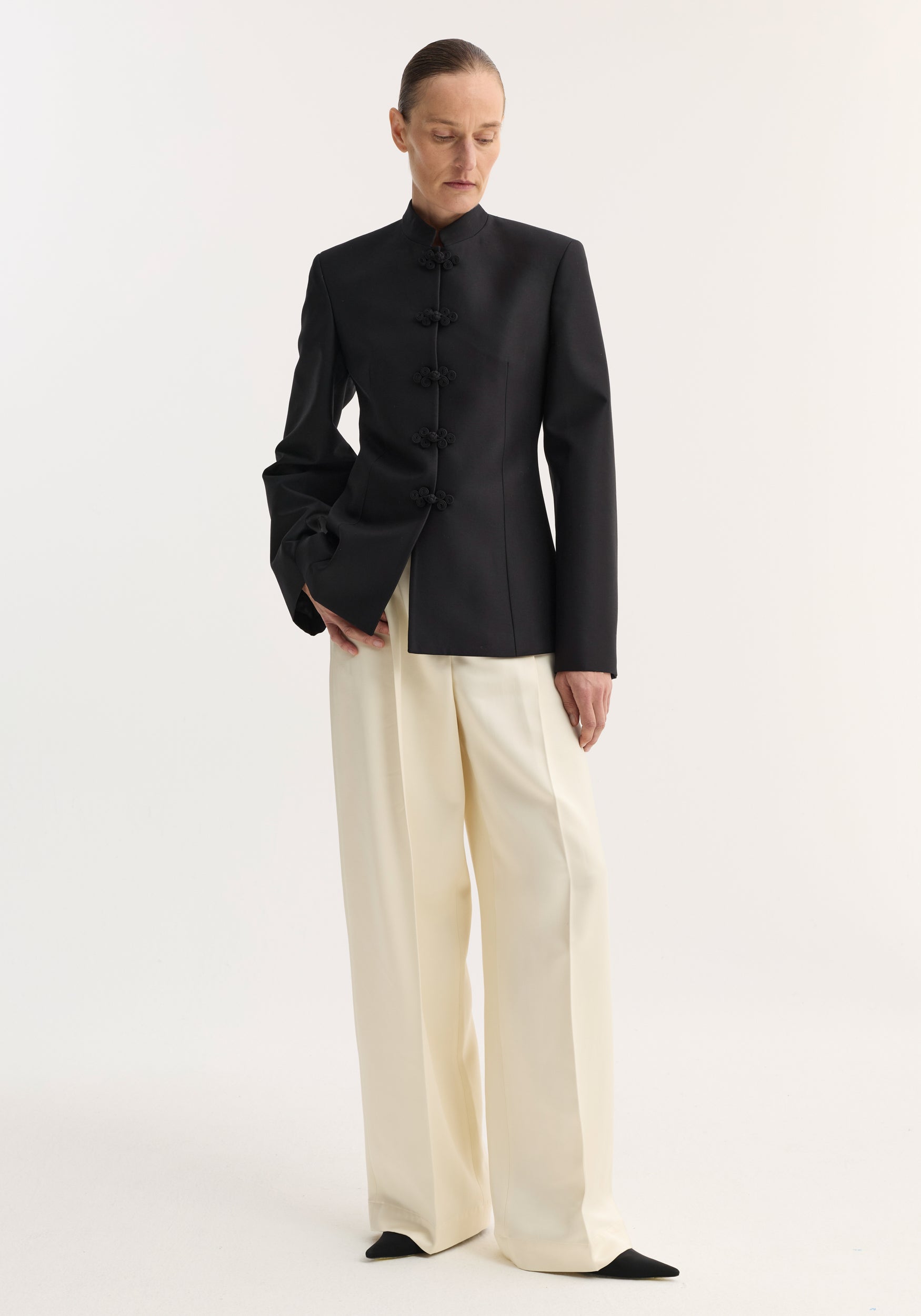 Woman wearing a black Sculptural pankou closure blazer | black with a stand collar and traditional pankou knot buttons, paired with high-waisted cream wide-leg trousers and black pointed-toe shoes.