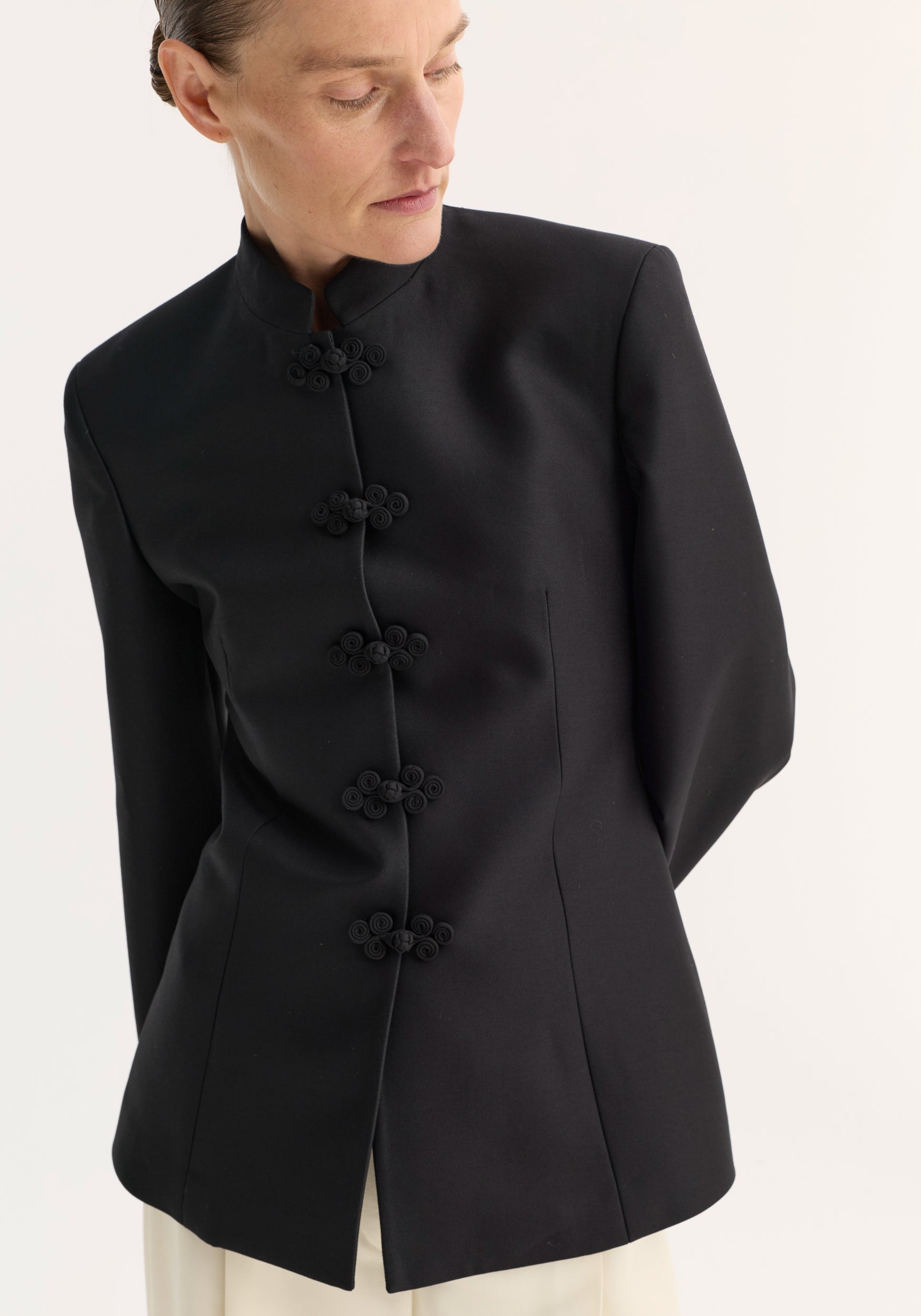 Woman wearing a black sculptural pankou closure blazer | black with a high collar and decorative knot buttons down the front, styled with a minimalist look.