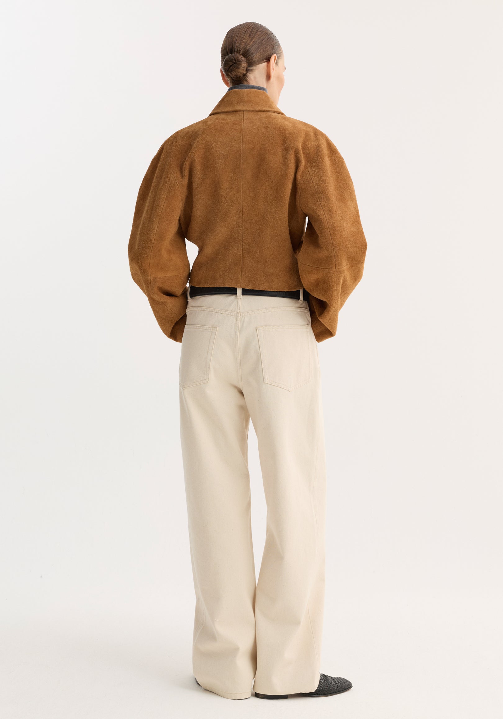 Rear view of a model wearing a rounded textured suede jacket | cognac in a warm brown shade paired with light beige wide-leg pants and black shoes against a plain white background.