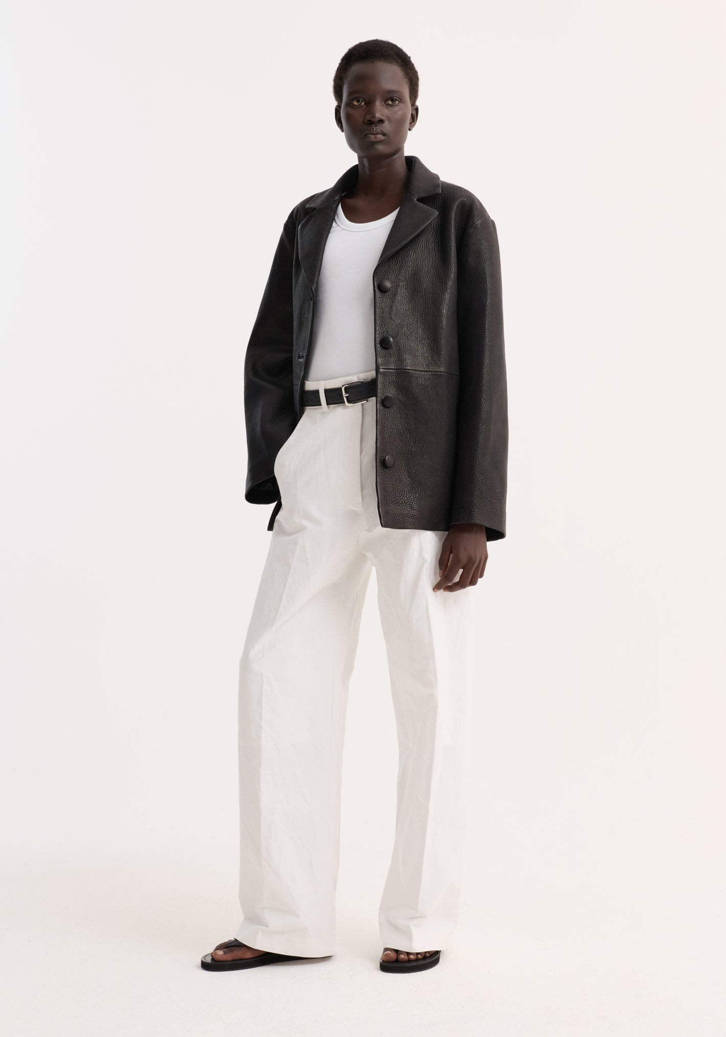 Model in a Leather jacket with small lapel | black, featuring a slightly oversized fit, button closures, and side seam pockets, paired with white trousers.