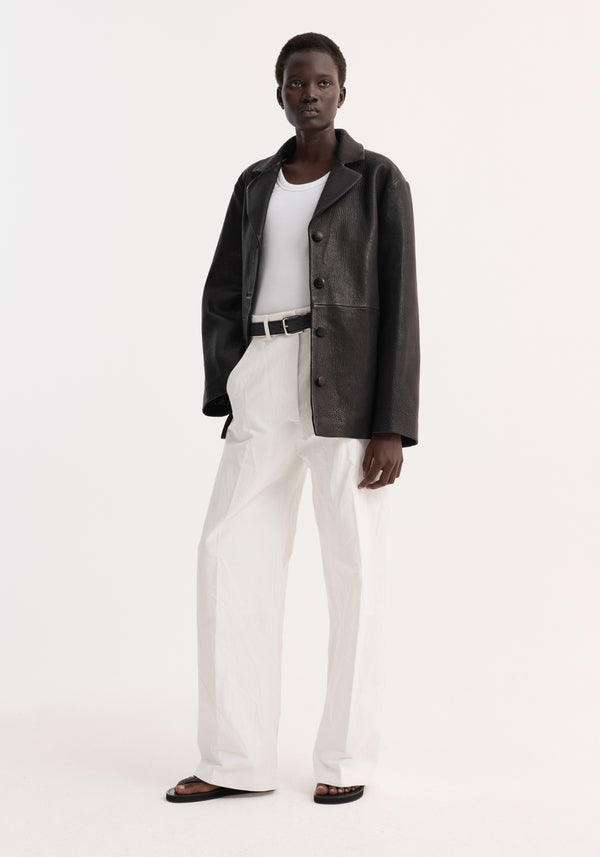 Model in a Leather jacket with small lapel | black, featuring a slightly oversized fit, button closures, and side seam pockets, paired with white trousers.