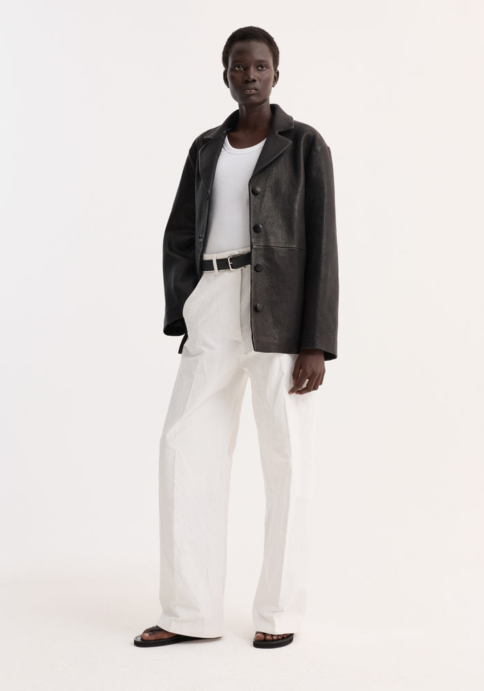 Model in a Leather jacket with small lapel | black, featuring a slightly oversized fit, button closures, and side seam pockets, paired with white trousers.