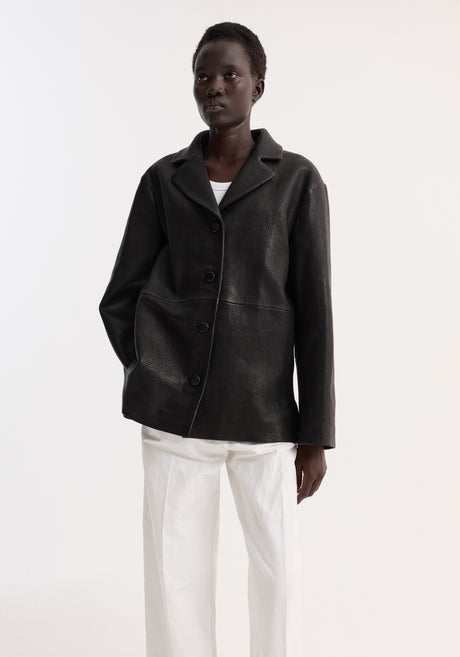 Model wearing a black leather jacket with small lapel, button closures, and side-seam pockets, over a white tank top and high-waisted trousers.