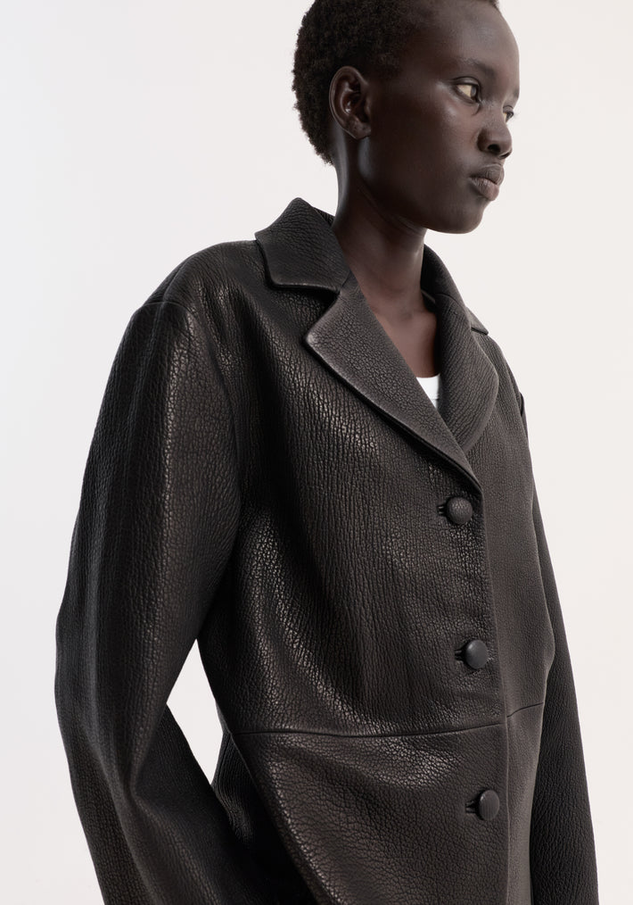 Close-up of a person wearing a structured black leather jacket with a small lapel, showcasing its boxy silhouette and three covered buttons.