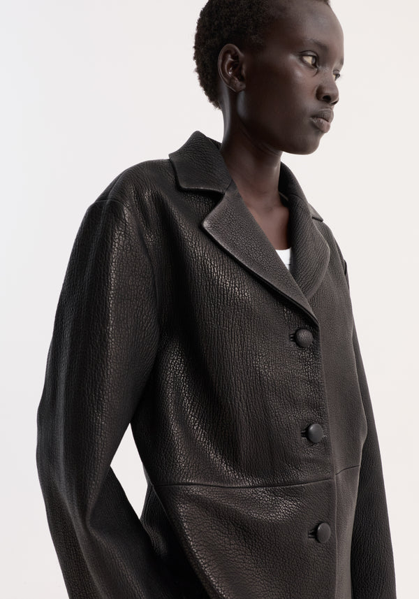 Person wearing a black Leather Jacket with Small Lapel, featuring a notched collar, large leather buttons, and textured heavy-grain leather, highlighting its structured, boxy silhouette.
