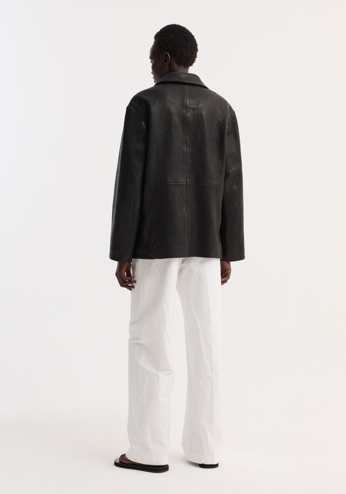 Person wearing a black Leather jacket with small lapel, showcasing its boxy cut and leather texture, paired with white trousers and black sandals.