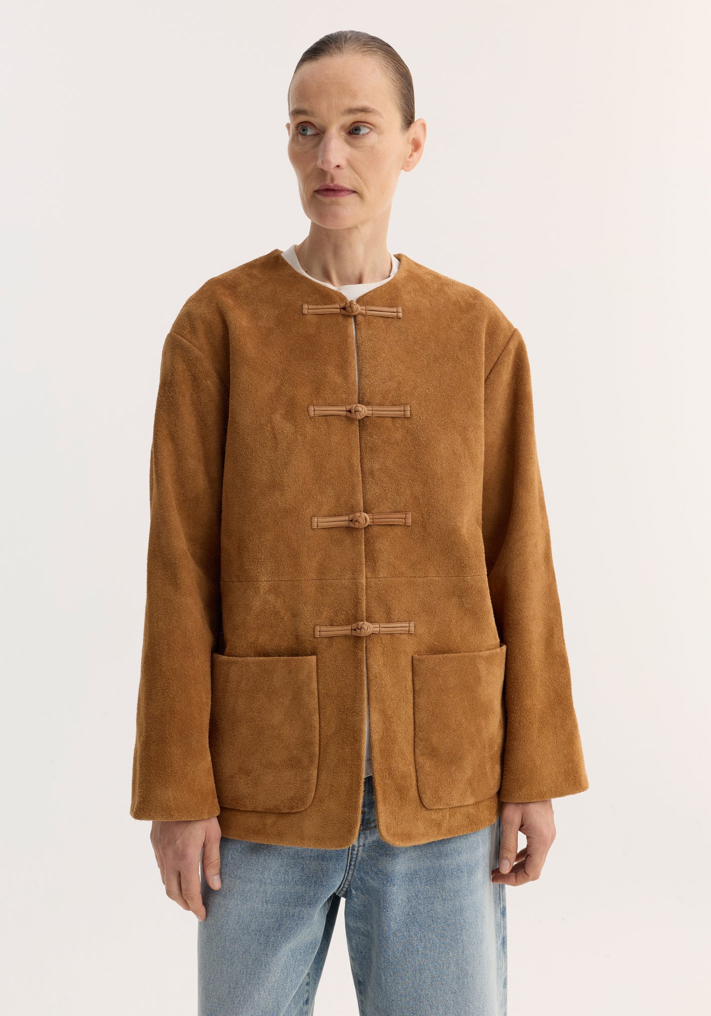 Pankou closure suede jacket in cognac with embroidered edges, featuring traditional toggle closures, patch pockets, worn over a white top and light-wash jeans.