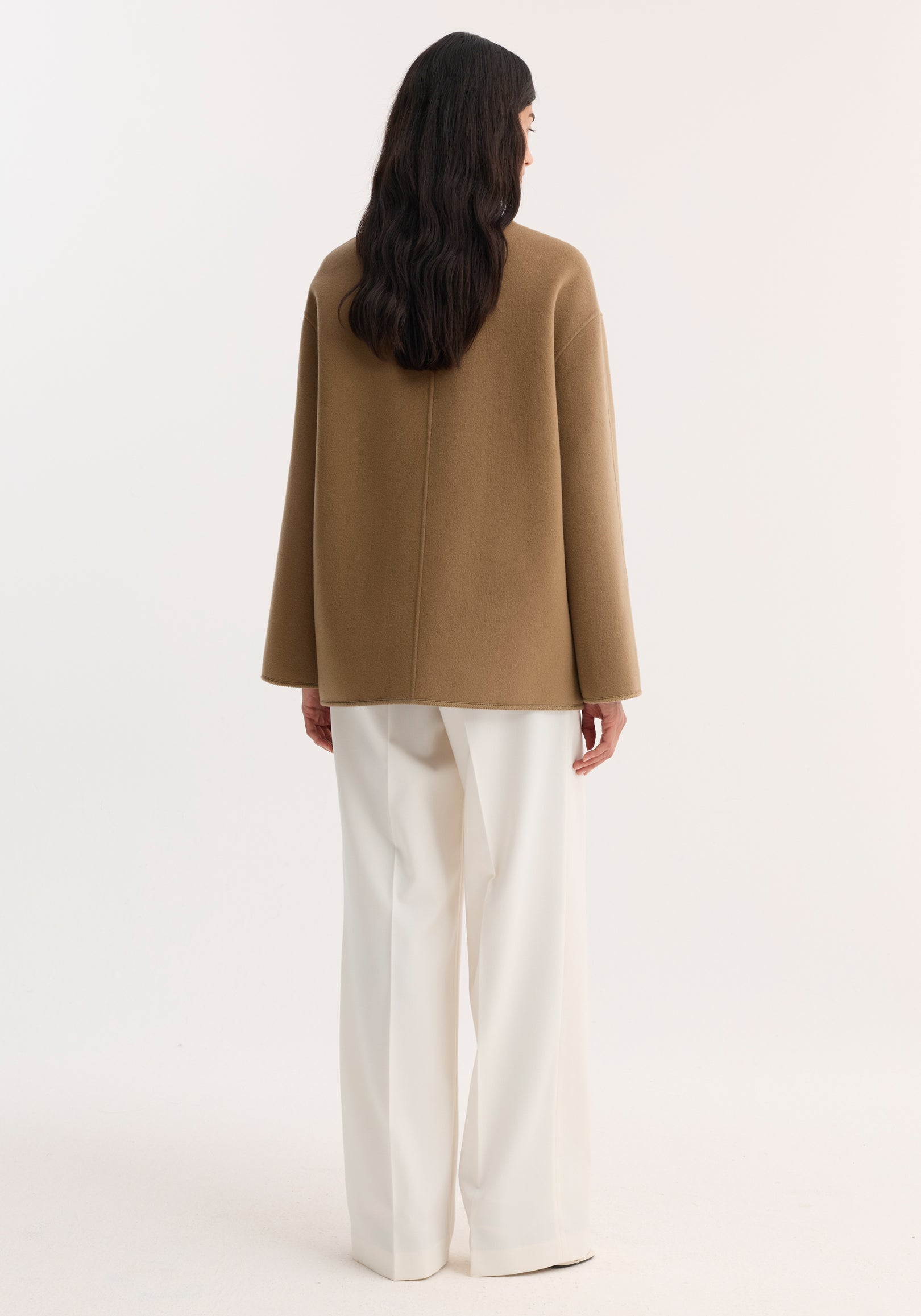Woman wearing a camel-colored double-faced wool pankou jacket viewed from the back, paired with loose white pants against a plain white background.