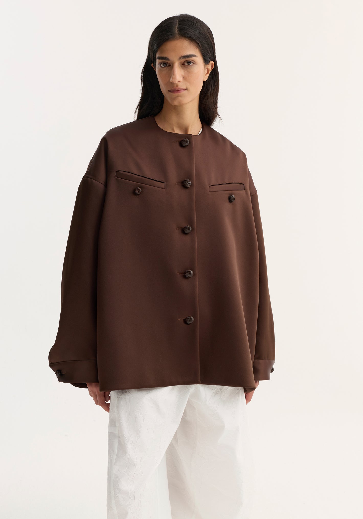 Model wears the Western heavy satin jacket in espresso, featuring oversized fit, leather-knotted buttons, chest welt pockets, and dropped shoulders, paired with wide-leg textured trousers.