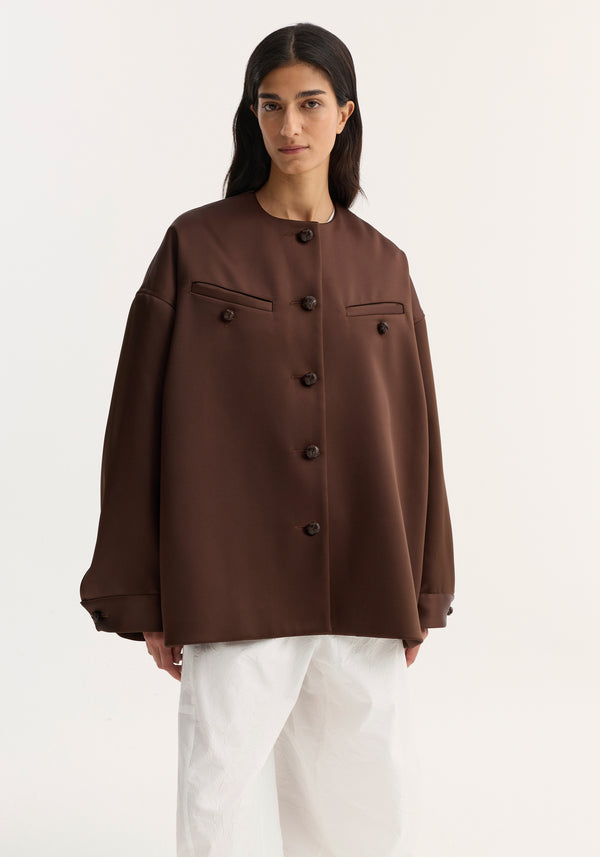 Model wears the Western heavy satin jacket in espresso, featuring oversized fit, leather-knotted buttons, chest welt pockets, and dropped shoulders, paired with wide-leg textured trousers.