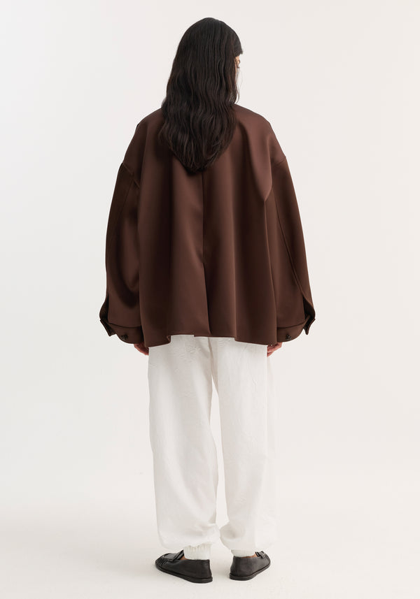 Back view of a model in an oversized Western heavy satin jacket with leather-knotted buttons and chest welt pockets, paired with white tapered trousers.