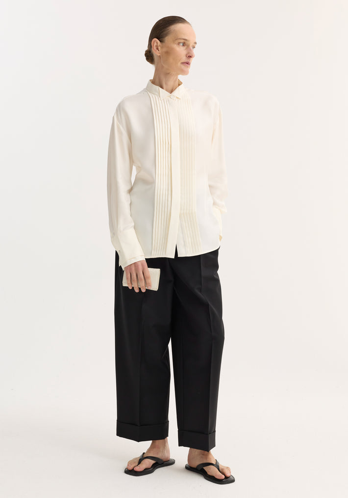 Woman wearing Shaped tuxedo silk shirt with stand-up collar, pleated front, and double cuffs, paired with black wide-leg trousers and thong sandals, holding a wallet.