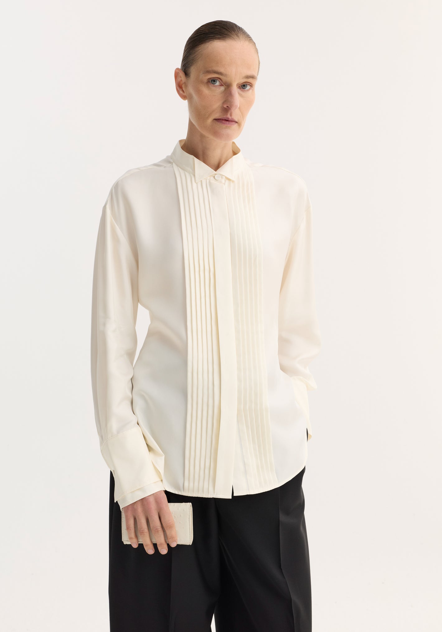 Shaped tuxedo silk shirt in cream on model, featuring a tuxedo-style pleated front, stand-up collar, double cuffs, paired with black trousers.
