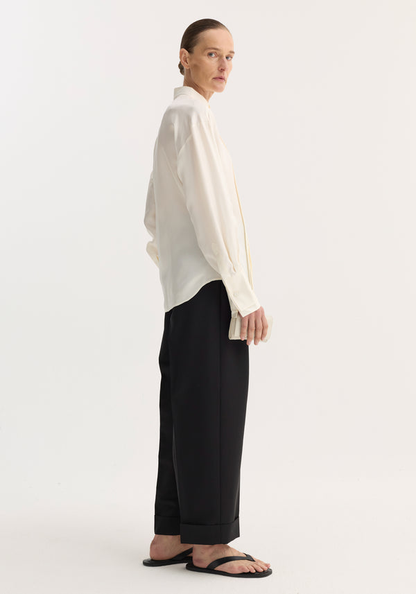 Shaped tuxedo silk shirt in cream with a stand-up collar, double cuffs, and pleated front, worn by a tall model holding a cardholder.