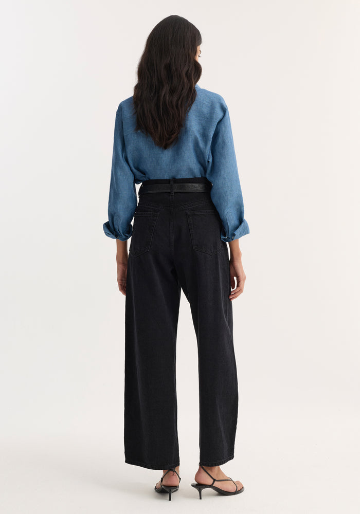 Woman modeling Barrel leg denim | black, showcasing high-waisted, wide-leg silhouette from the rear, paired with a light blue shirt and black strappy sandals.
