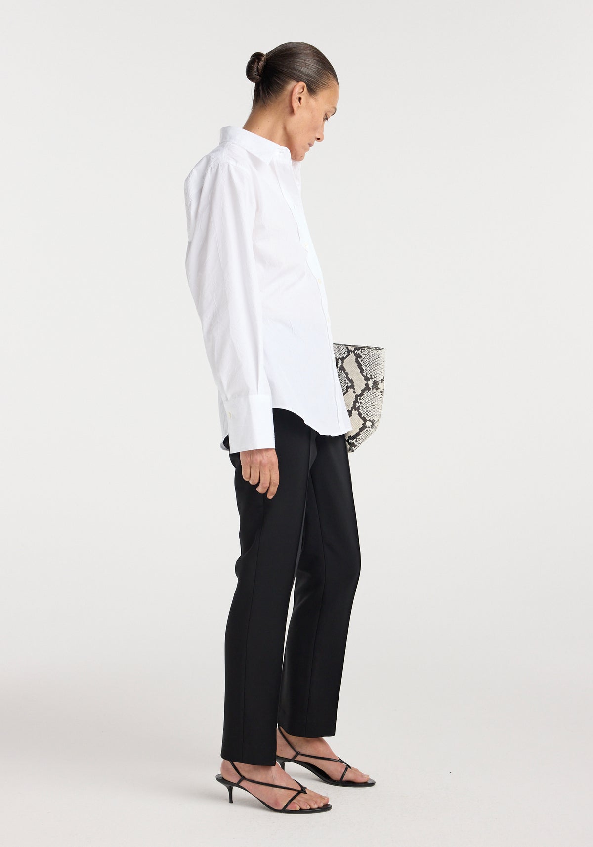 Woman wearing a tailored Shaped shirt | optic white paired with black fitted pants and strappy black heels, holding a snakeskin-patterned clutch against a plain background.